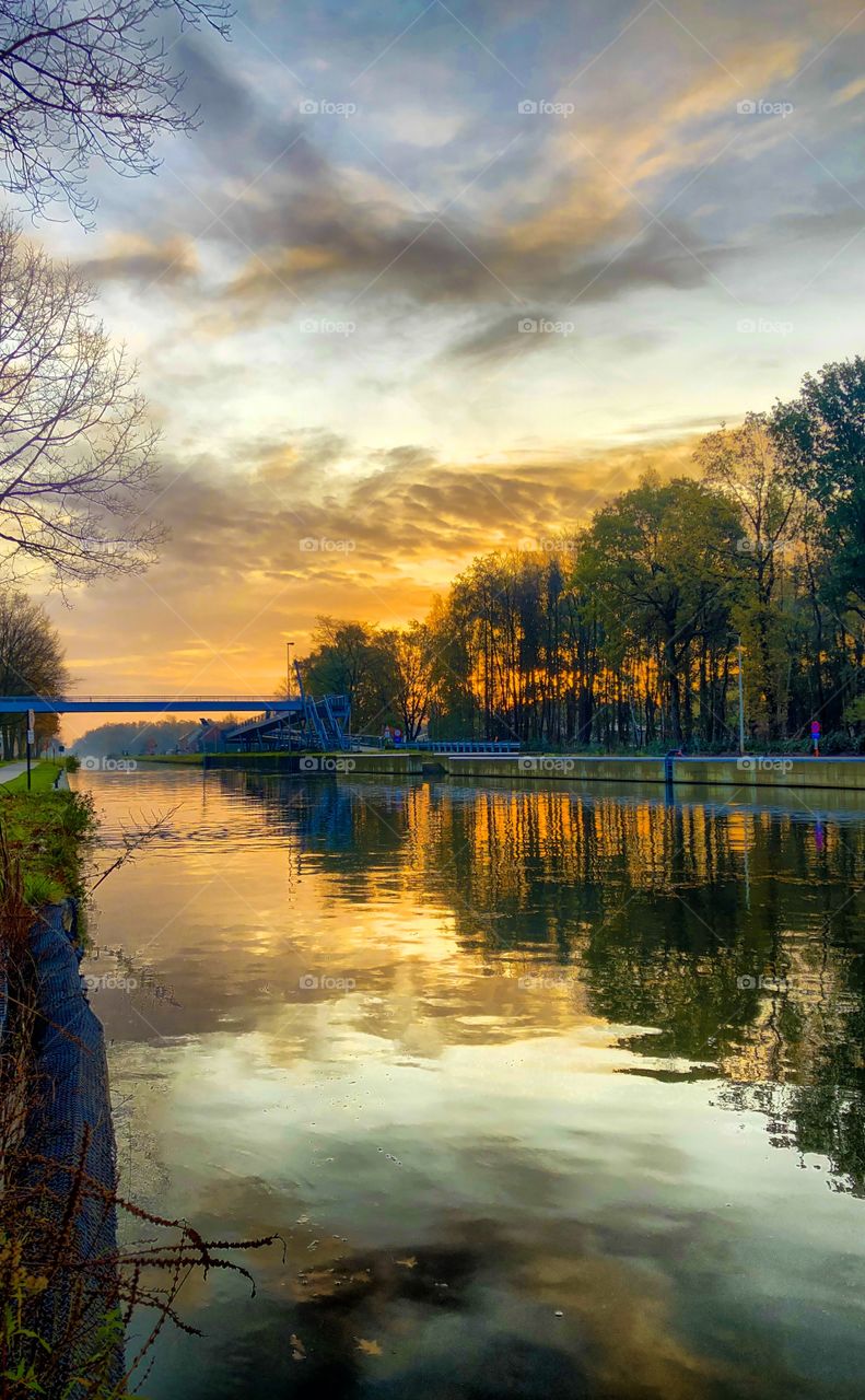 Dramatic and colorful sunrise or sunset sky over a canal of river running through a forest landscape, reflected on the surface of the water