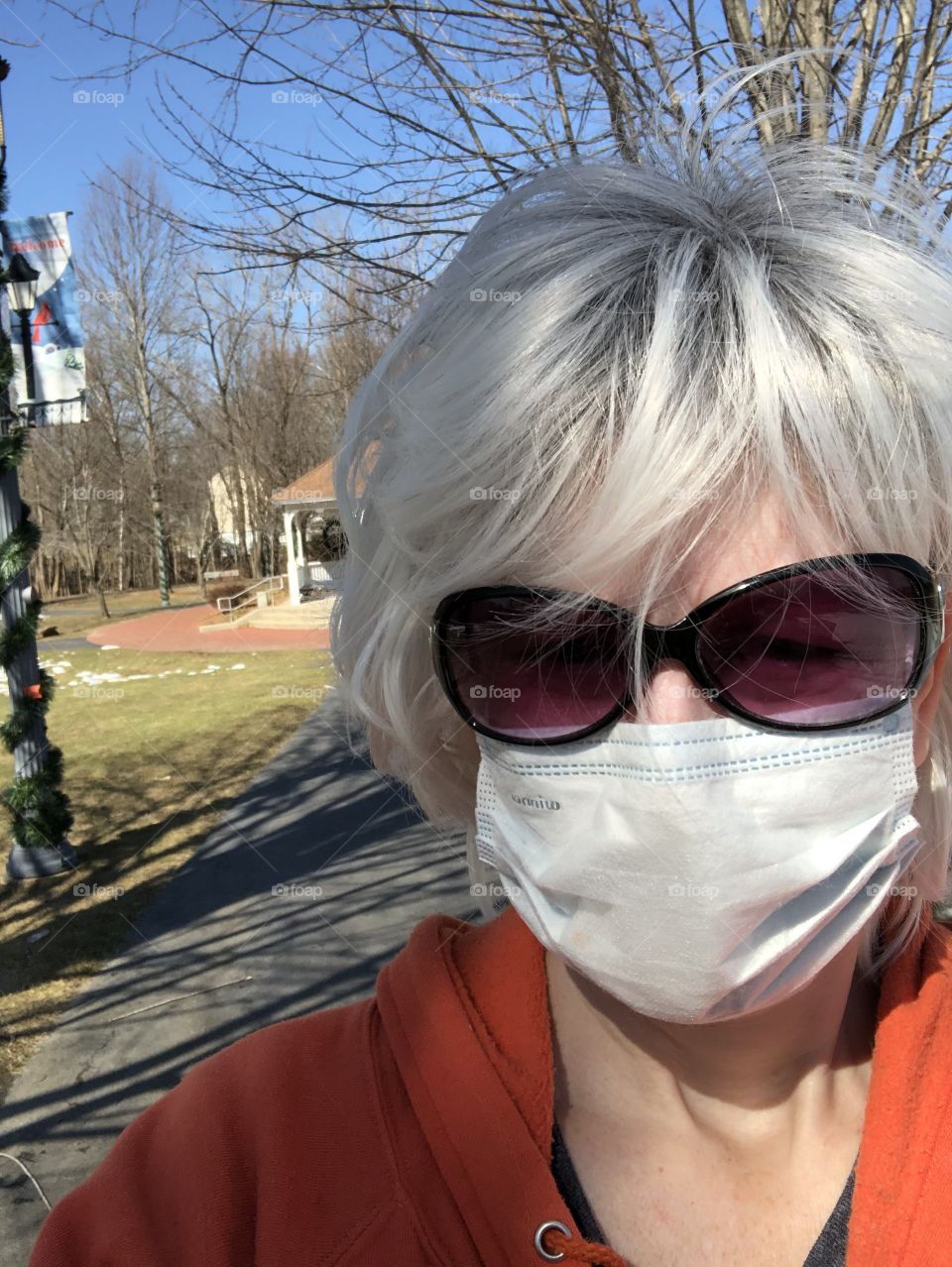 Woman with blonde hair and sunglasses is wearing a white face mask and standing alone outside at a park on a mild, sunny day in March. 