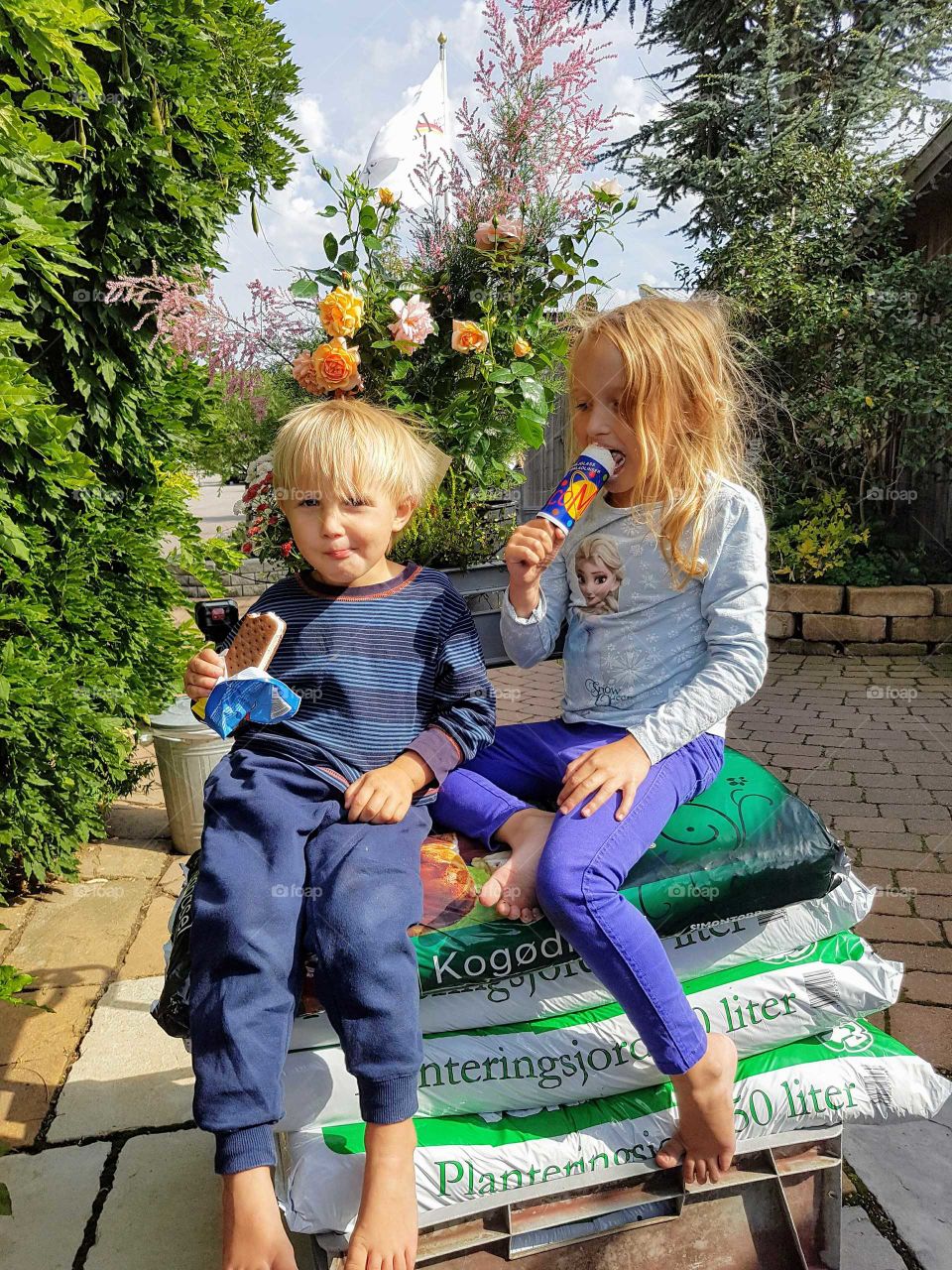 two kids on a barrow, sitting on piles of soil with flowers in background