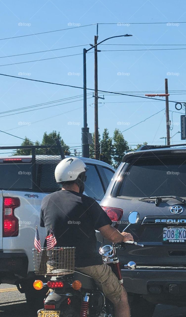 man wearing a white helmet riding a motorcycle with small American flags attached to the back in Oregon traffic