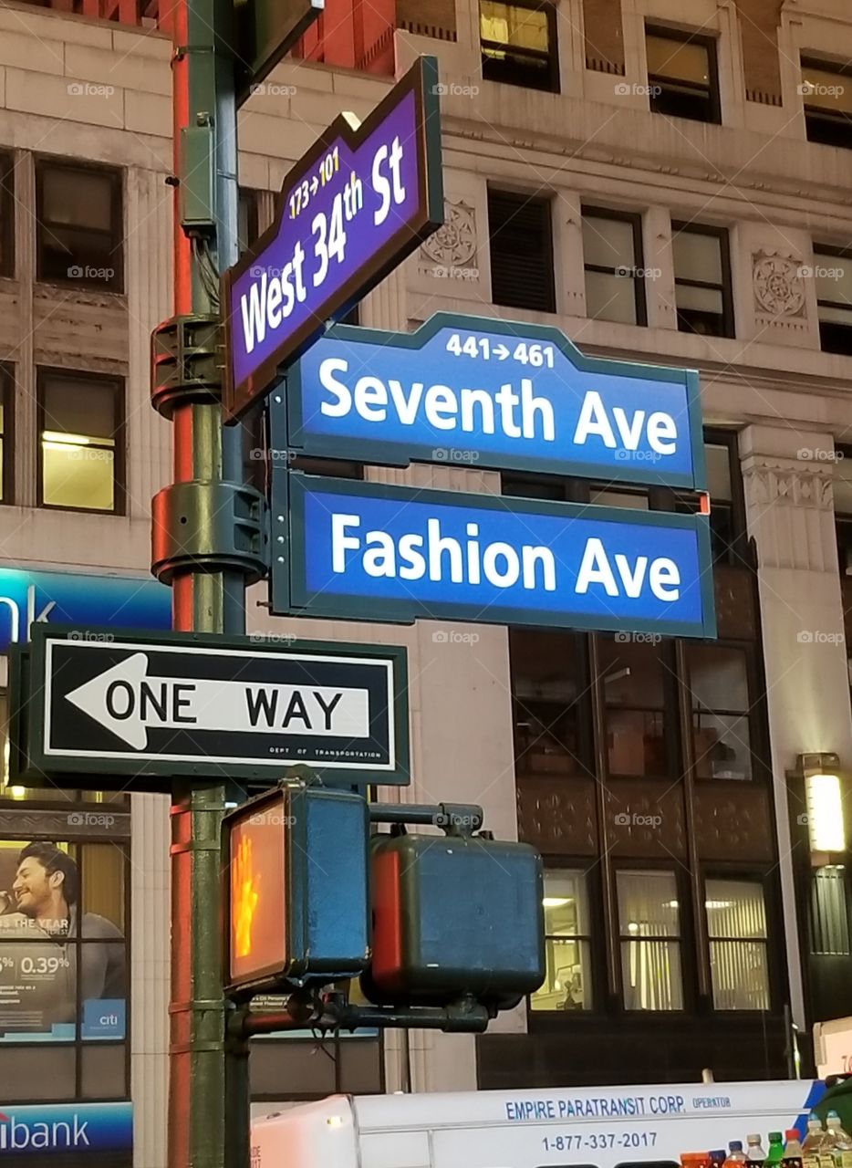 New York City street signs in vivid color. The signs are on a corner located in downtown Manhattan. The signs are blue, and purple.  This is the corner of West 34th Street, Seveth Avenue and Fashion Avenue near Macy's. The photo was taken at night.