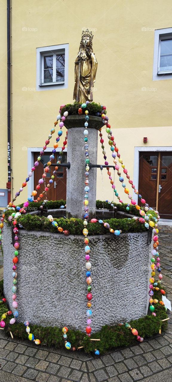 This fountain in the German city of “Regensburg” is festively decorated for “Easter” using a green reef and multicolored eggs on a string. The gold figurine on top is part of the permanent decor. 2023. Hypnotic Productions