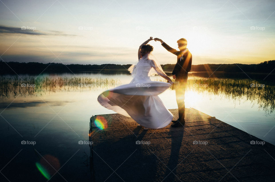The groom holds bride's hand on the lake shore at sunset. Wedding concept