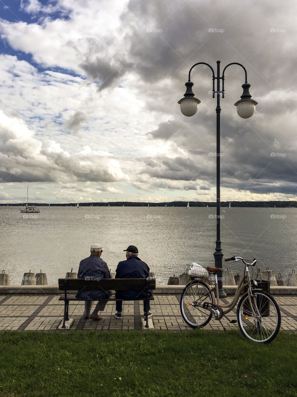 A view of two old men from behind sitting on a bench with parked bicycle next to the bench in front of a lake in a sunny day.