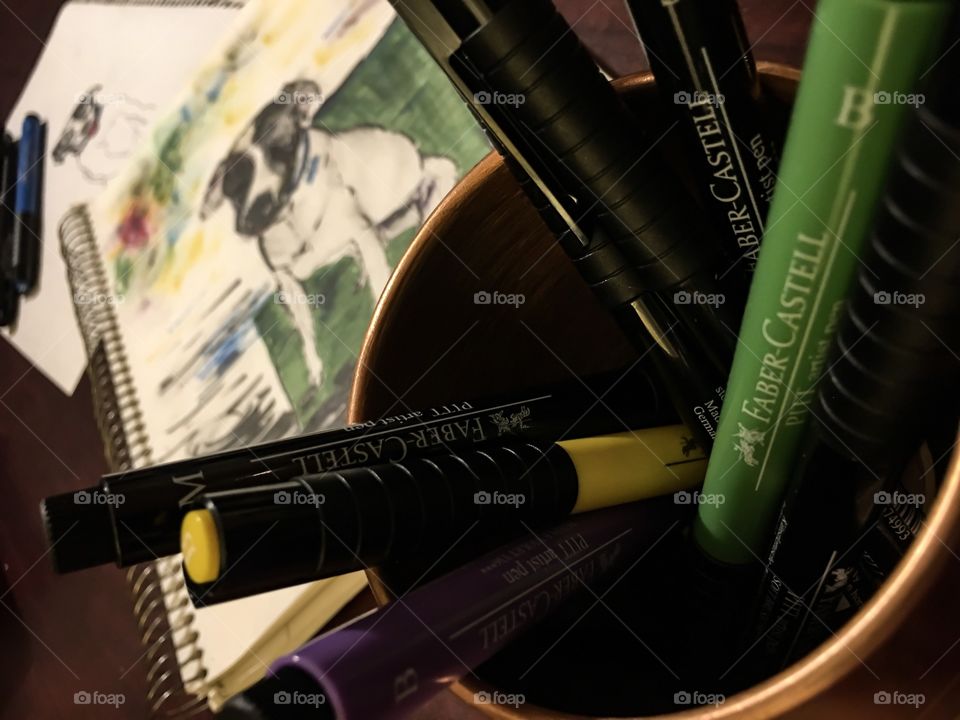 Faber-Castell Pitt artist pens in copper mug with colorful portrait of family pet dog on artist sketchbook in background using color techniques of the PITT artist pen’s various capabilities 