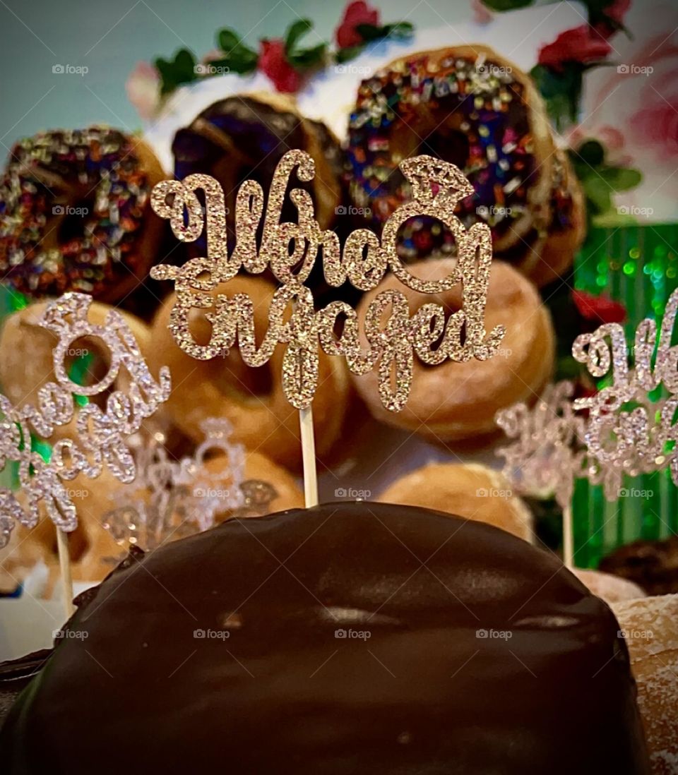 A Boston crème donut in a donut bar party with a toothpick glitter sign stating “we’re engaged”