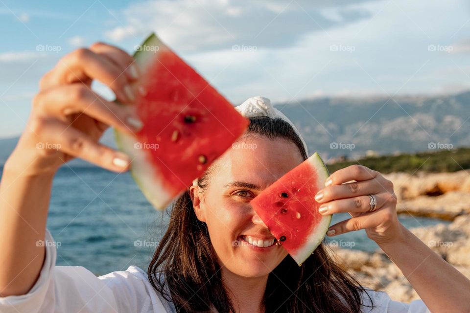 Portrait of pretty girl standing on beach, holding pieces of watermelon in front of her face