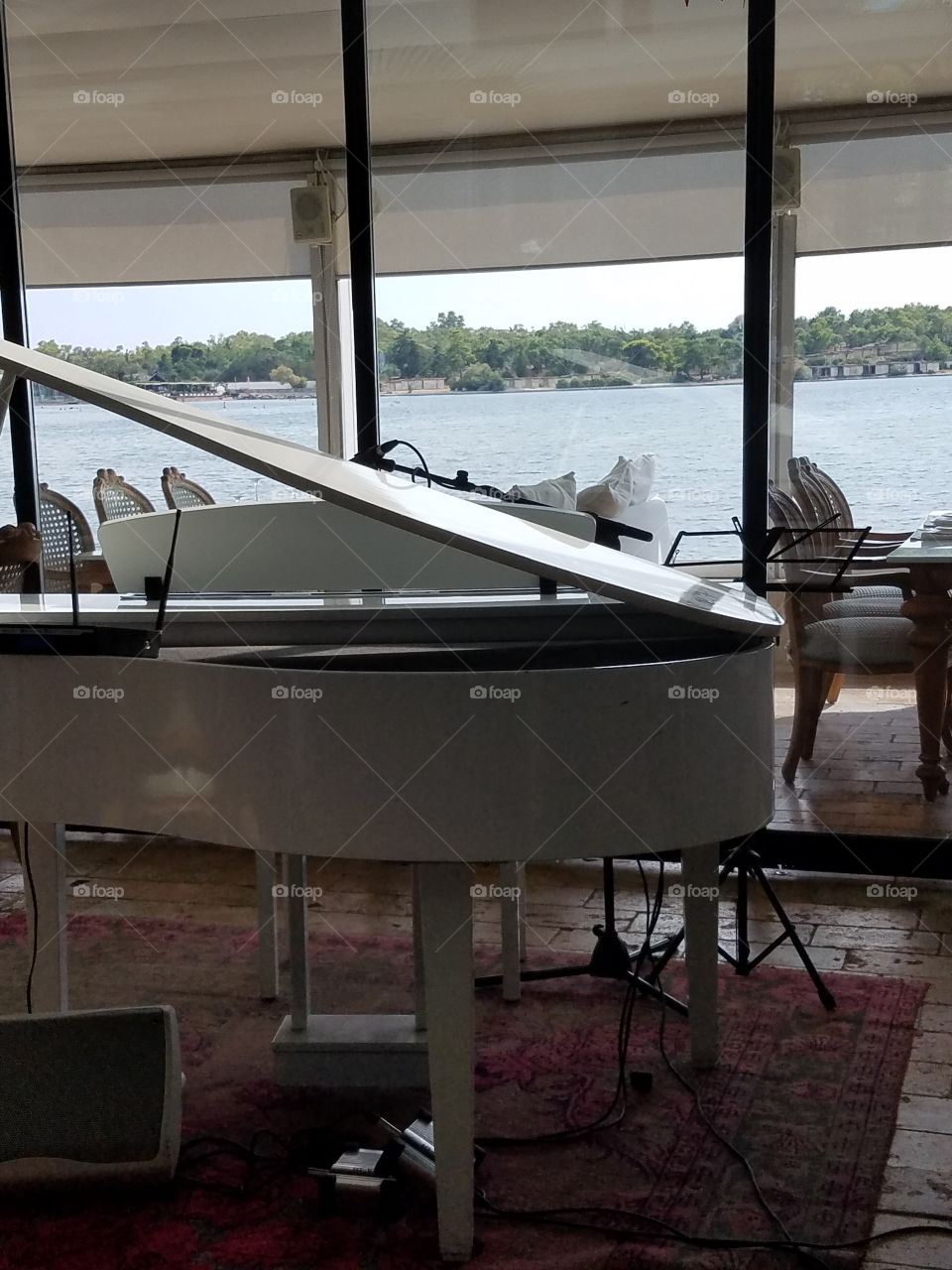 A white piano with a background of a window displaying the ocean and green trees. The piano is in an elegant restaurant in Greece.