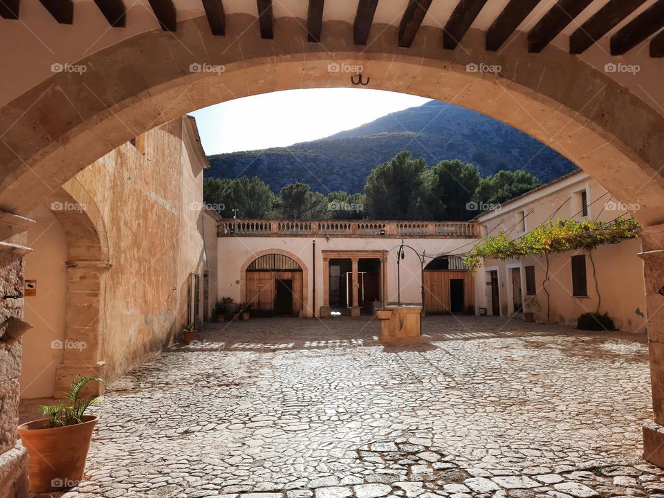 Majorcan traditional manor patio with stone floor and a well. In the foreground a traditional stone arch. Vegetation and mountains at the background. Calvia, Majorca, Spain.