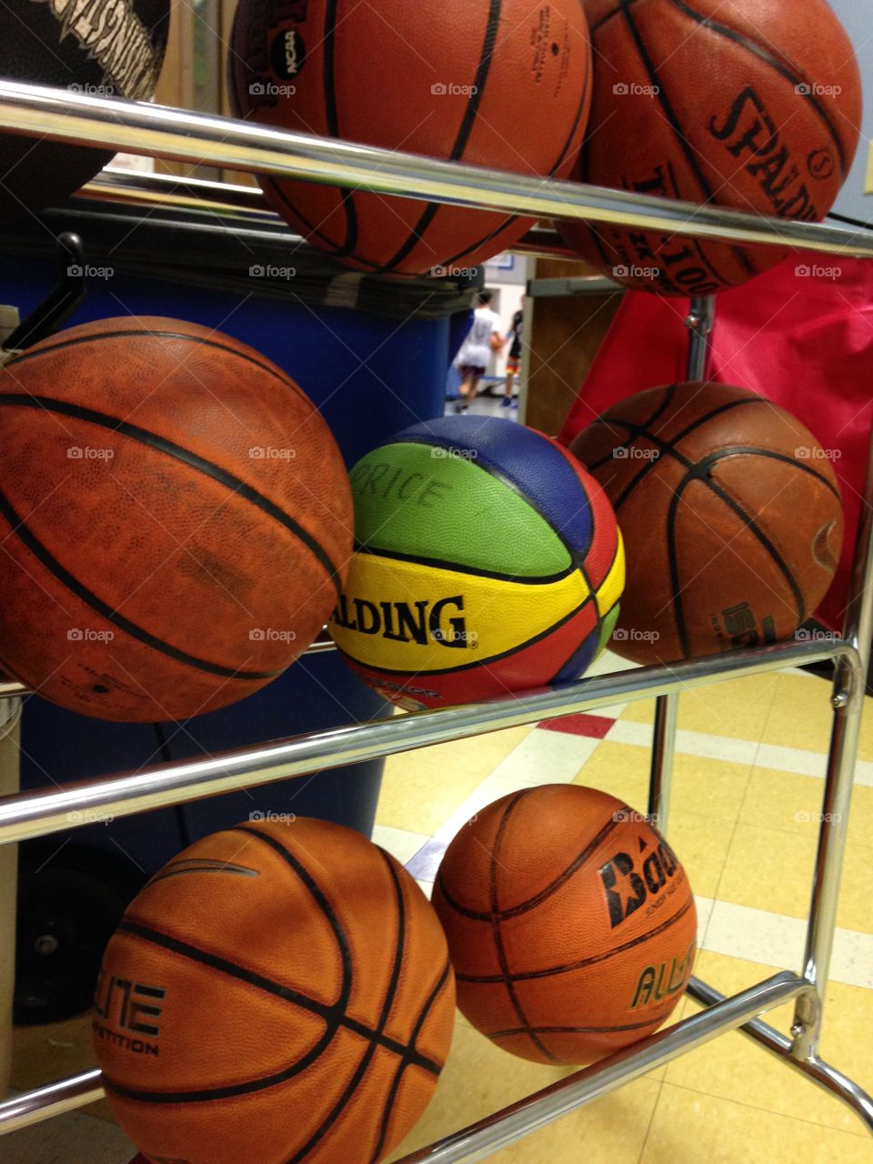 Basketballs on a rack. People visible behind in the distance playing basketball.
