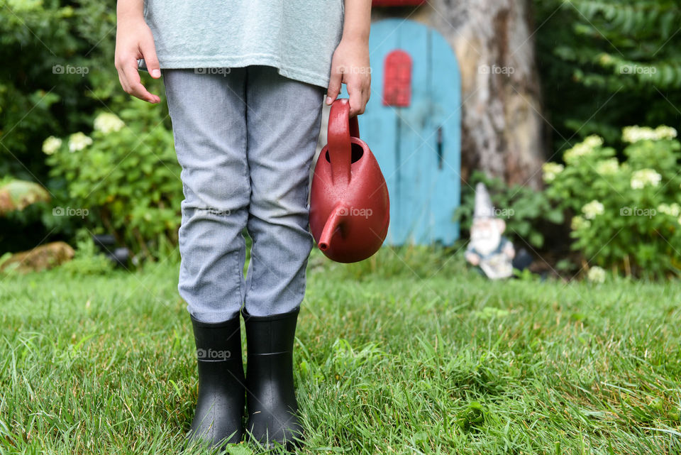 Bottom half of a child holding a red watering can in front of a little gnome garden outdoors