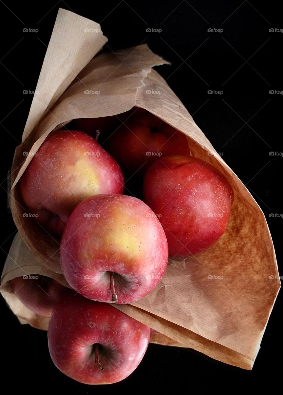 On a mirrored surface lies paper rolled up into a cone containing red apples.  One apple has rolled out and is reflected in the mirror surface.  Black background