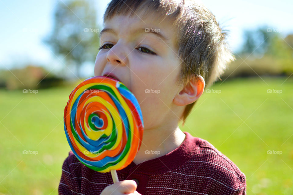 Toddler boy eating a large rainbow colored lollipop outdoors