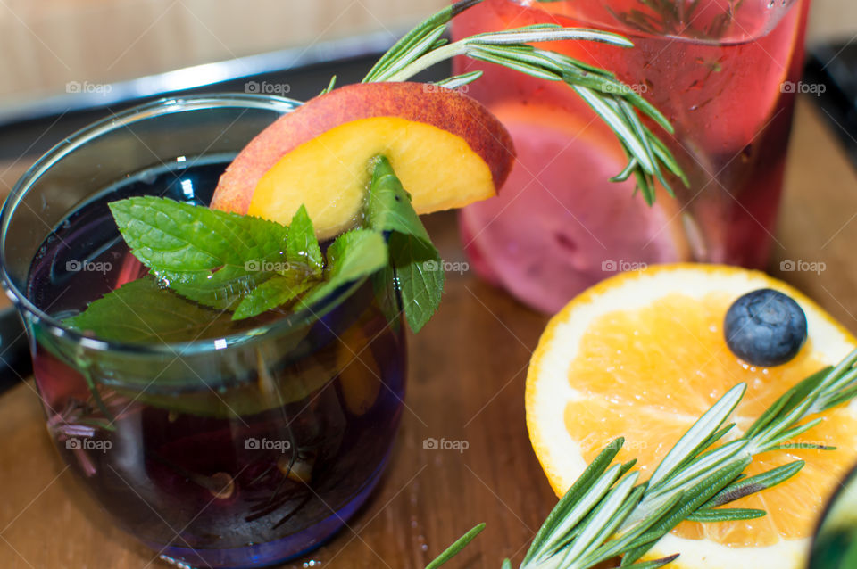 Glass of homemade peach, blueberry, orange, cherry fragrant juice garnished with fresh garden rosemary, peach slice and mint leafs on dark wood conceptual healthy drink citrus flavored water photography 