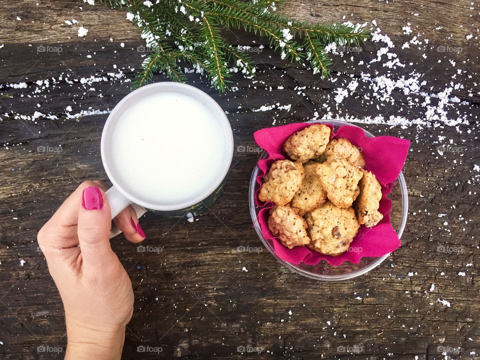 Woman hand wearing pink nail polish holding a mug of milk with cookies beside on wooden table powdered with snow