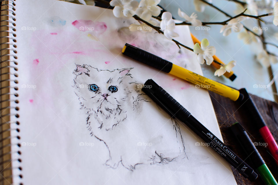 Artist workshop spring table with flowers and Faber-Castell PITT artists pens on sketchbook with sketch of white kitten made using smudging, water (watercolor) and sketch techniques of the PITT pens tranquility and creativity conceptual photography 