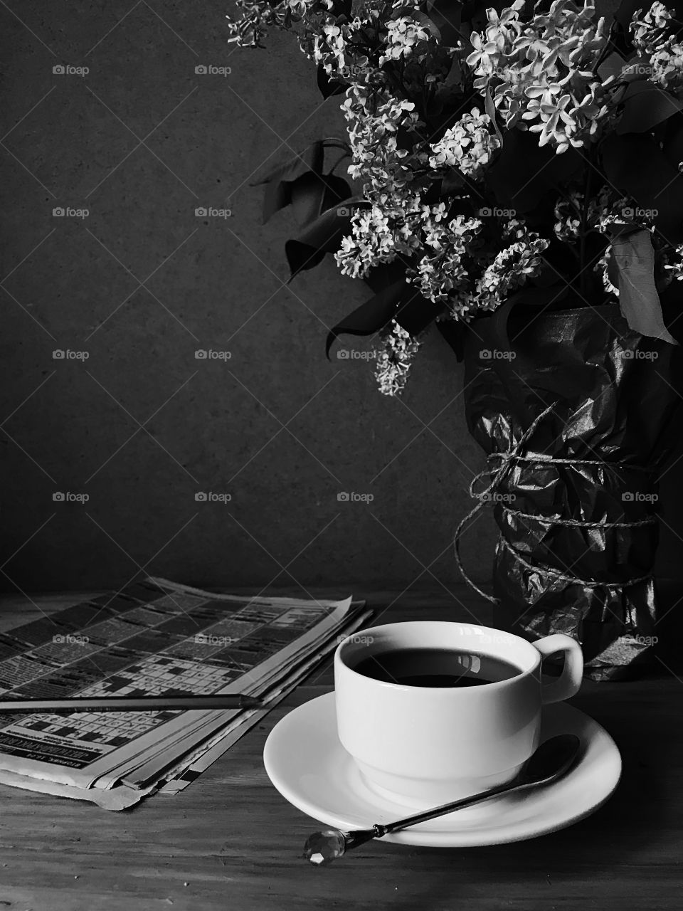 Cup of coffee, newspaper with crossword and pencil, bouquet of lilac flowers on rustic wooden background black and white 