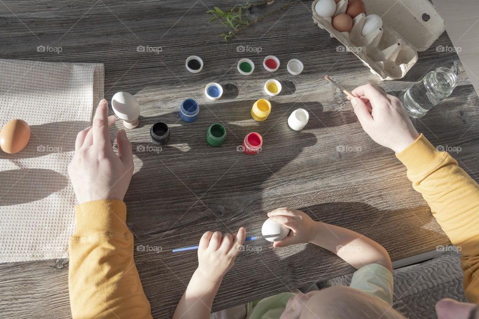 A little son and his father together in their free time paint Easter, chicken eggs with paints and brushes for the holiday.  A child and a man create a craft and natural product in their family business.