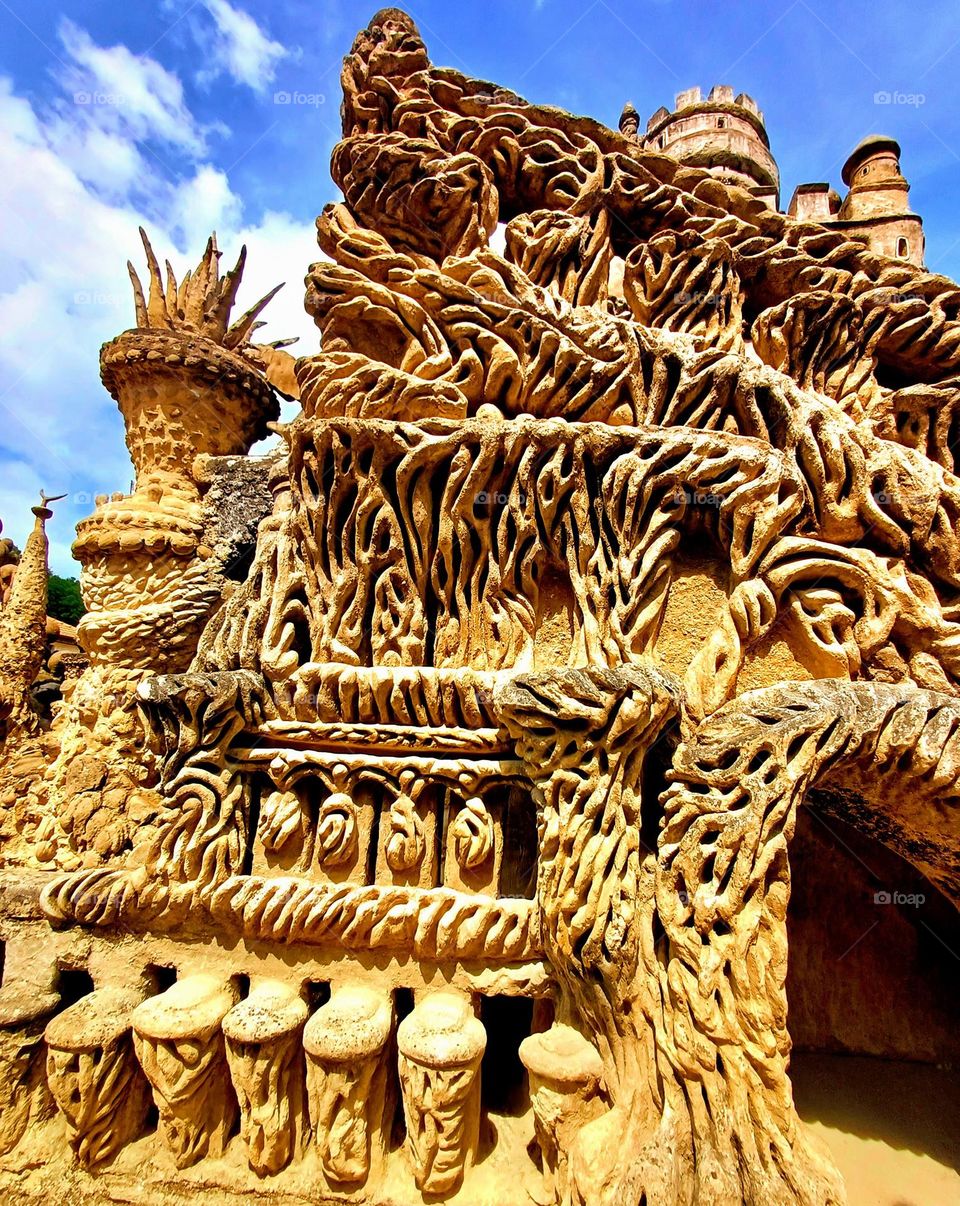 It's a section of the Palais Idéal du Facteur Cheval, showcasing its intricate and imaginative stonework. The structure is adorned with a variety of elaborate patterns and textures, resembling natural formations like twisted roots or vines.