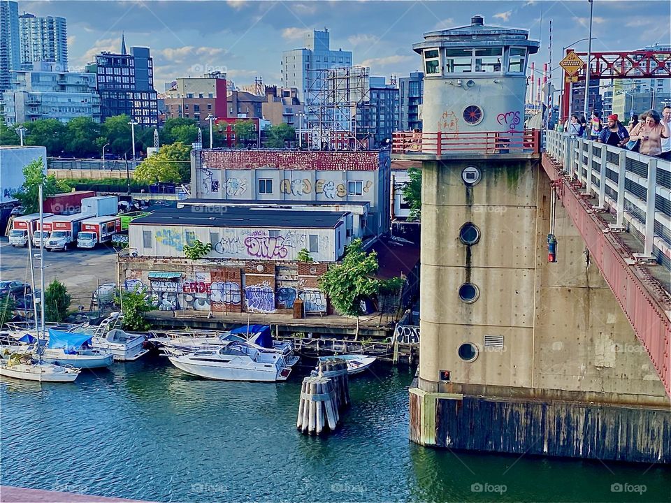 A graffiti style rendition of “Long Island City” at “Newtown Creek” seen from the “Pulaski Bridge”. One of its main support columns is on the left with a tower cabin on top. The wood pilings mark the position of the boats. 2022. Hypnotic Productions