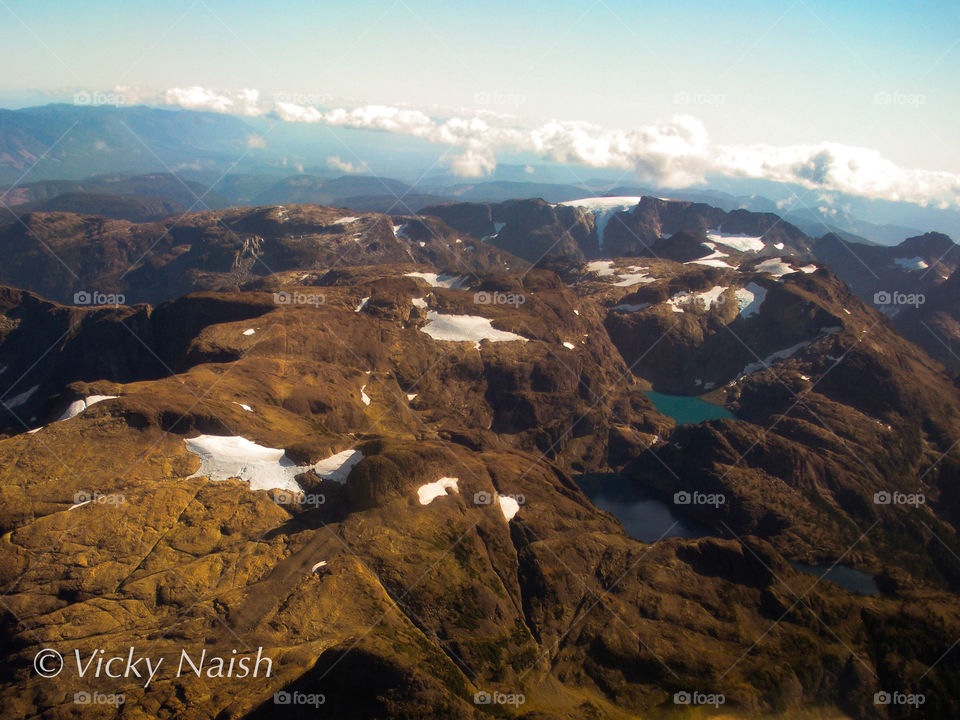 A beautiful view of summer in the island mountains seen from a low flying aircraft. Lower worn warmer mountains in the foreground & towering craggy snow covered peaks behind. 