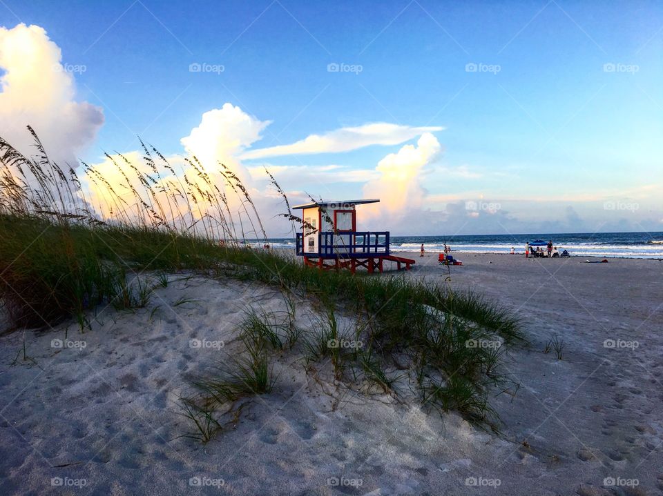 Beach scene showing a sand dune with grass and some plants as well as a lifeguard stand and the ocean in the background. The start of the sunset is seen in the blue sky. Photo taken near Cocoa Beach, Florida 