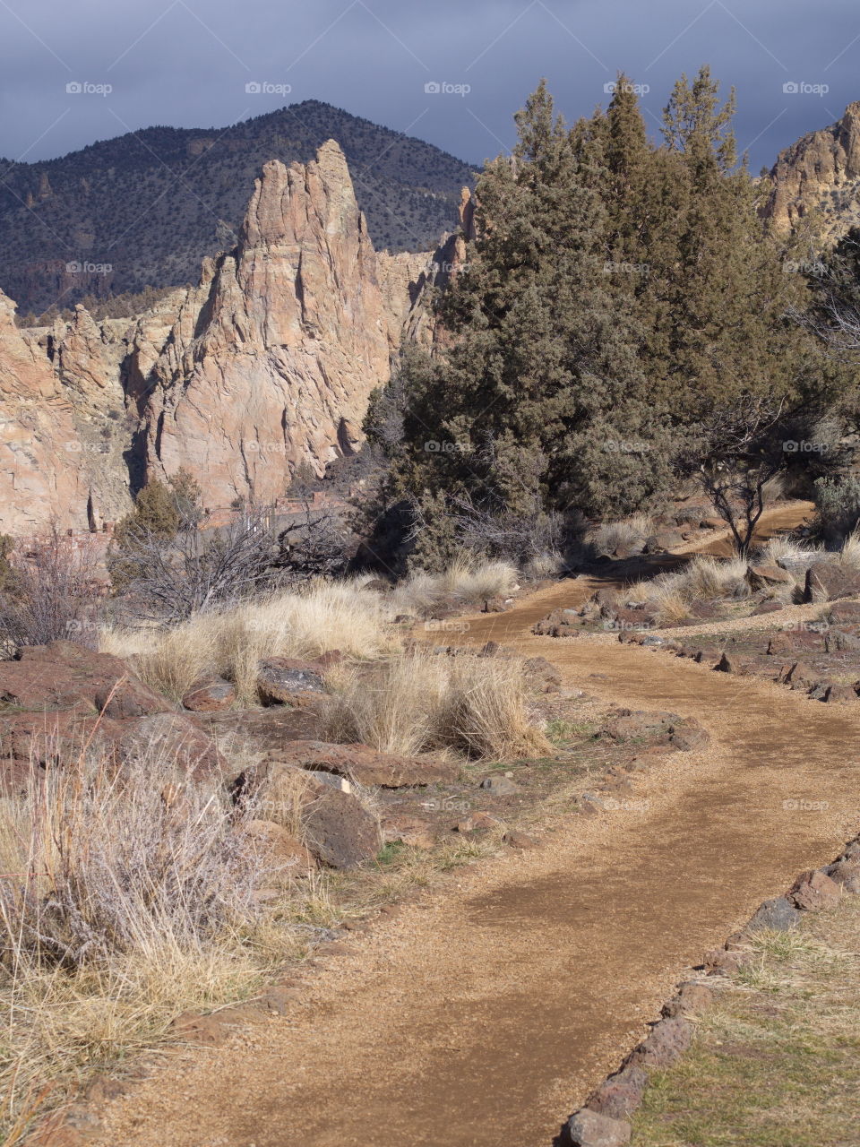 A pathway winds through the rocks, juniper trees, and wild grasses leading to the jagged cliffs at Smith Rocks State Park in Central Oregon with a stormy sky developing.