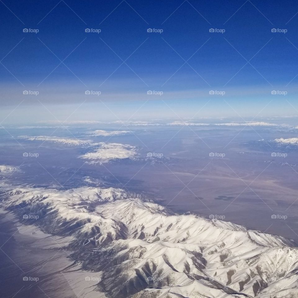 airplane view of snow covered mountain peaks with clouds and blue sky