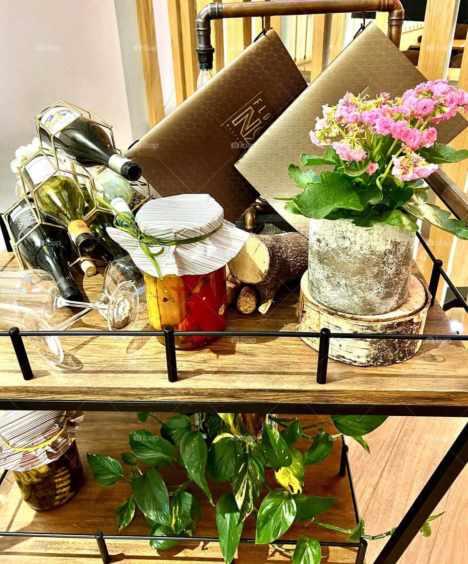 The photo shows a cozy composition: a pot with bright flowers, next to it stands a jar of pickles and bottles of wine, creating an atmosphere of home comfort.