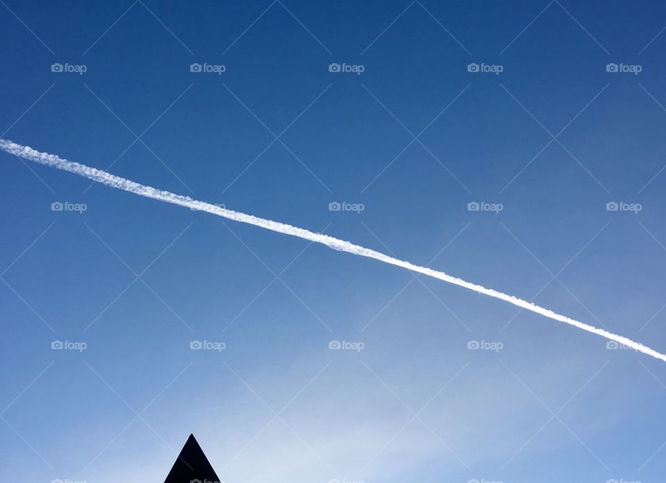 White jet stream across vivid blue sky, pointed rooftop at base of image, conceptual, minimalist, blue sky negative space 