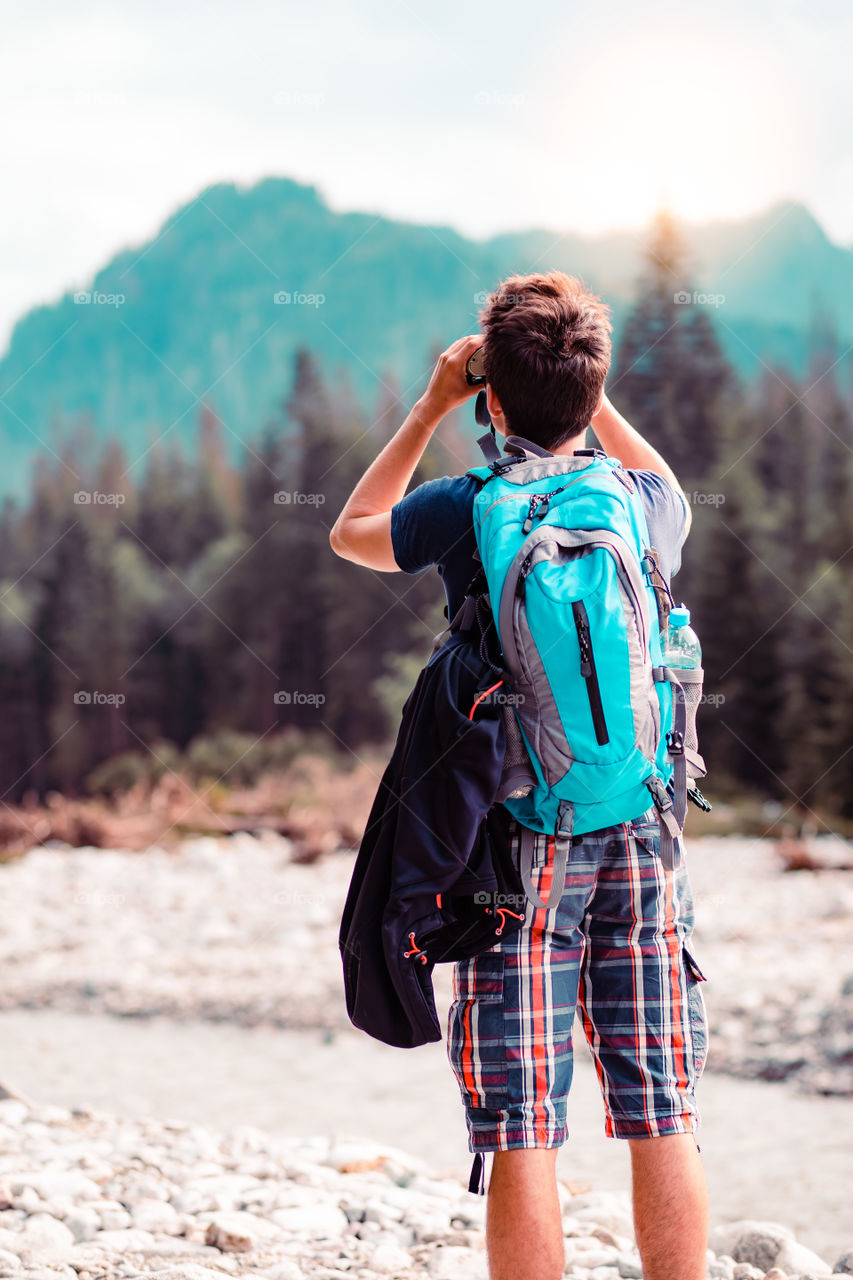 Young wanderer with backpack looks through a binoculars on mountains peaks, stands over a river. Boy spends a vacation in mountains, wandering with backpack, he is wearing sports summer clothes