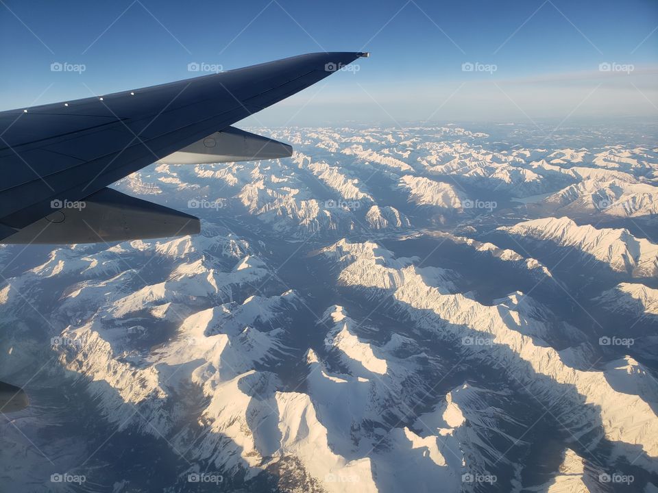 View from above, looking out of airplane window while flying over snow covered Canadian rockies during sunny day.