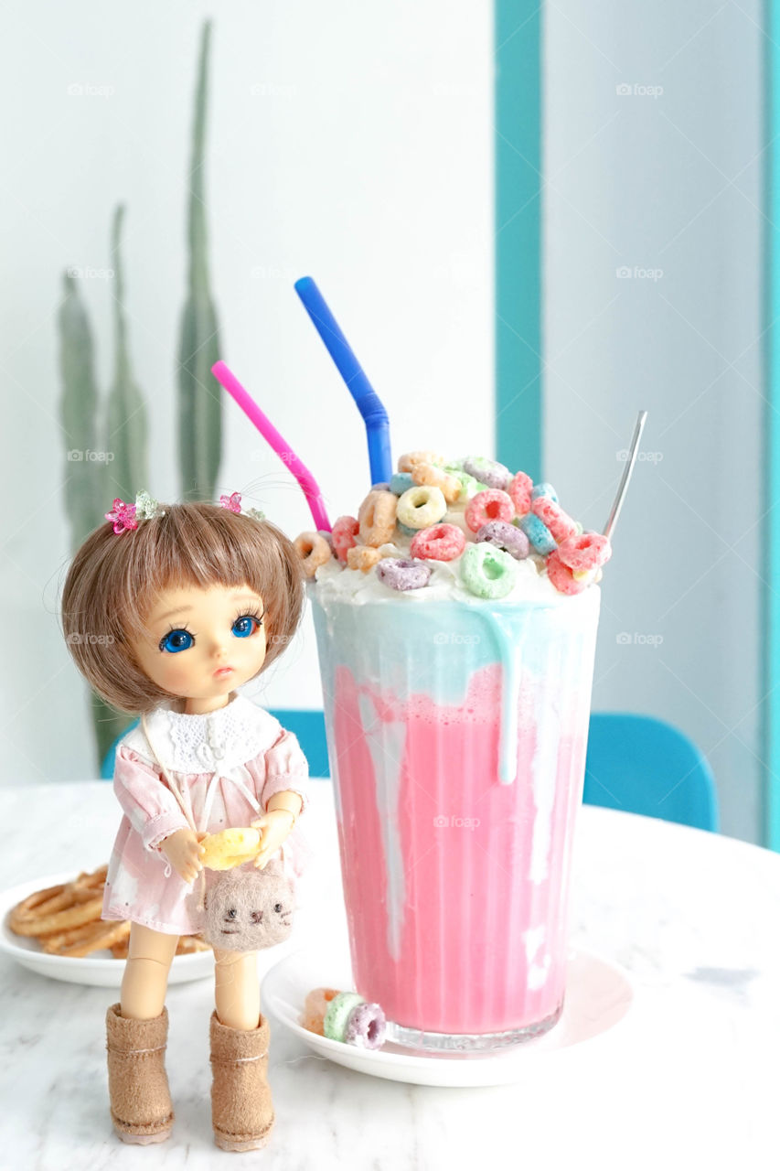 cute baby girl doll is holding a yellow cornflake in her hands. Rainbow milkshake is behind the little girl. Cute and sweet concept.