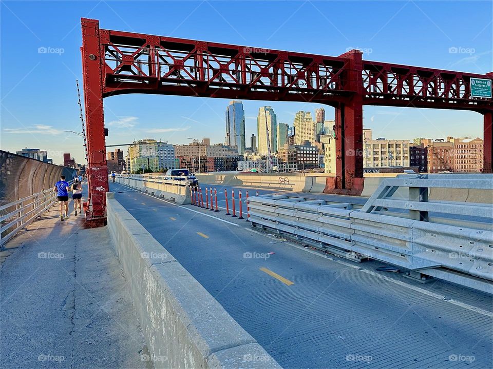 These are the pedestrian and bicycle lanes of the “Pulaski Bridge” at “Newtown Creek” connecting “Greenpoint”, Bklyn to LIC in the direction we are heading. LIC high rises bask in the golden glow of the evening sun. 2024. Hypnotic Productions