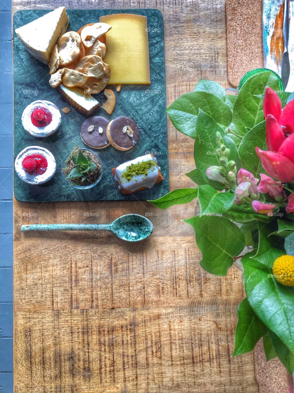 Food platter with sweet treats and cheese on wooden table with floral bouquet in corner. Copy space at bottom left