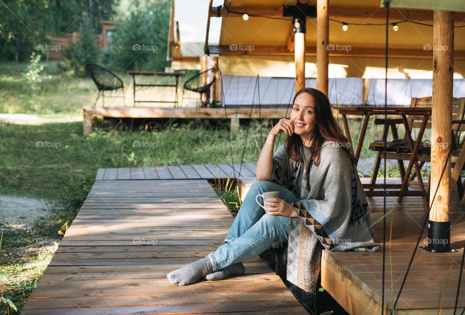 Young brunette woman in poncho drinking tea and relaxing in glamping in nature