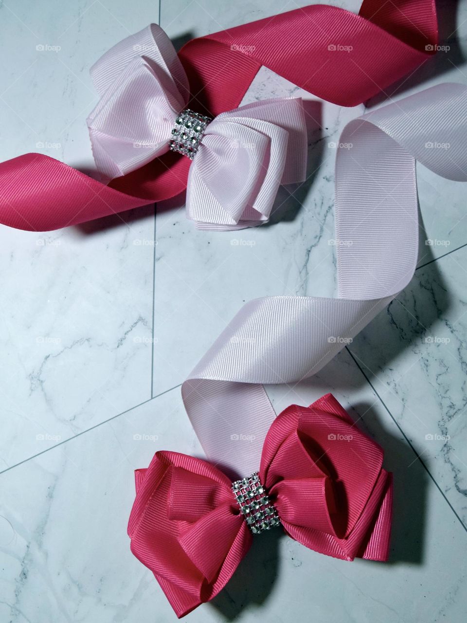 A baby pink bow secured with silver diamanté’s sits on top of curling hot pink ribbon, accompanied by a bright pink bow secured with silver diamanté’s sitting on top of curling pale pink ribbon on a clear white marble flatlay background.