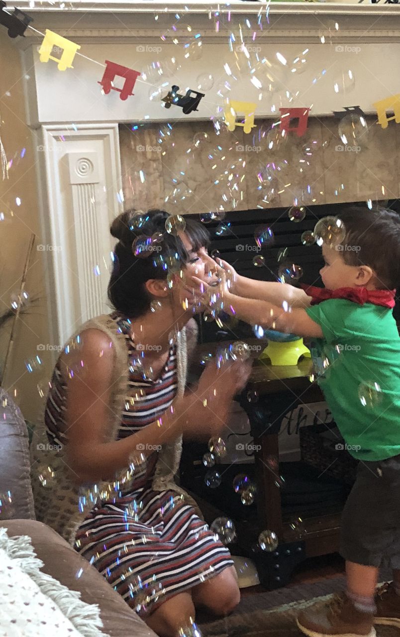 A bevy of bubbles surrounds this Mom and her two year old , arms stretched towards his mommas face at his birthday party! 