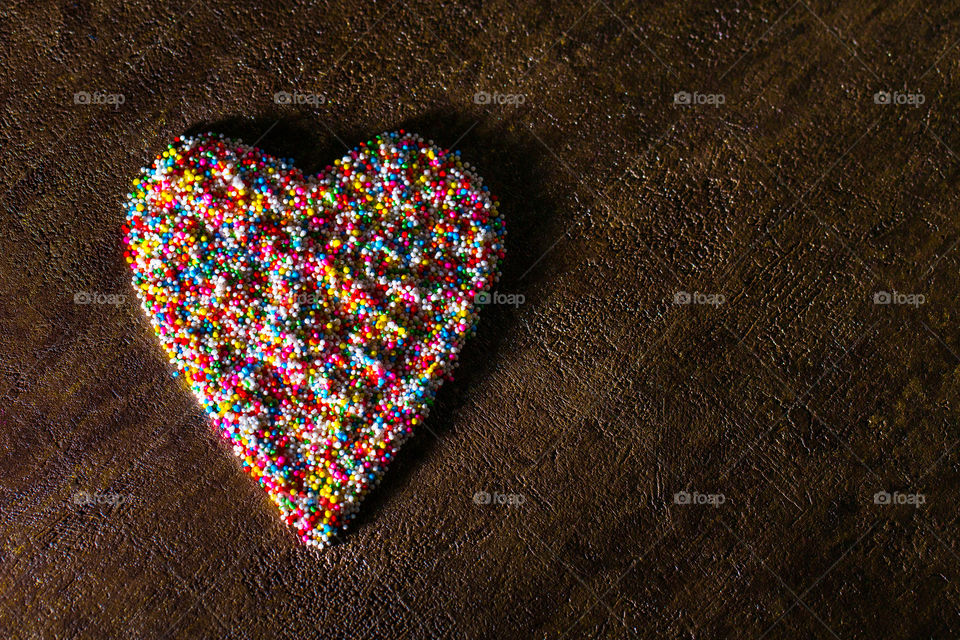 Heart shaped with ice cream and cake sprinkles using different colors. Dark textured background. Flat lay of heart. Side lighting with empty space on right.