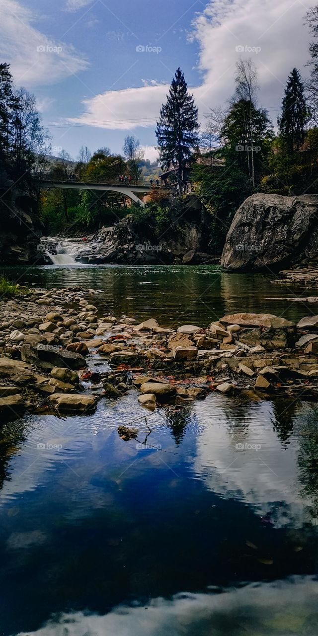 Green blue water of the mountain river, sky and treetops reflection in the water. People on the bridge look on the waterfall afar. Stone river's shore, rocks, forest and mountains. Vertical waterscape, tourists visit beautiful nature.