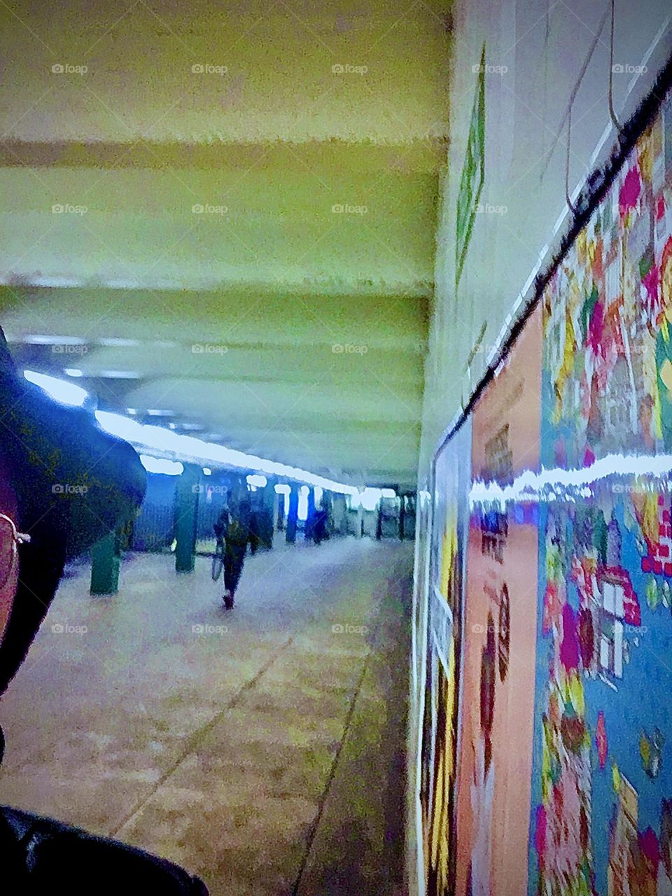 What looks like outer space is the „Van Alst Av 21st St“ „G“ train station in LIC, Queens with neon lights going off into the distance, commuters walking in winter clothes and a colorful art poster displayed on the wall. 2020. Hypnotic Productions
