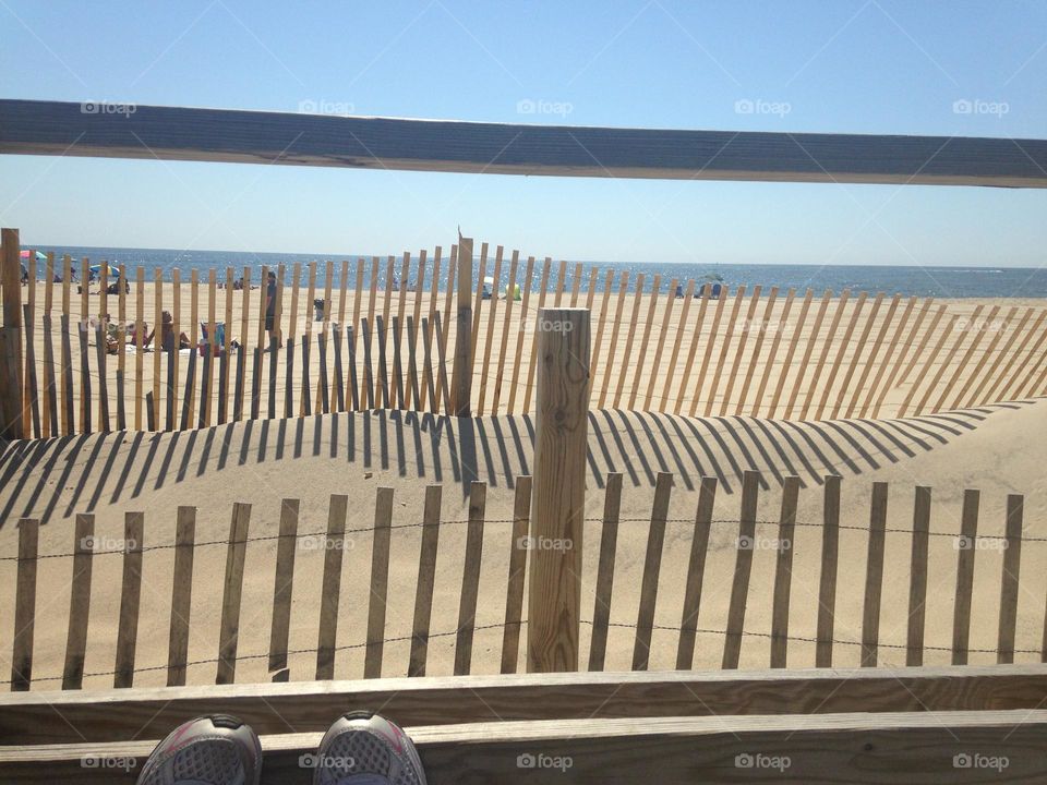 A photo of the beach from a bench on the Point Pleasant Beach boardwalk in NJ. The tips of my sneakers rest against the bottom of the boardwalk fence, the ocean and beach goers are in the distance, and the beach fence makes an interesting pattern.