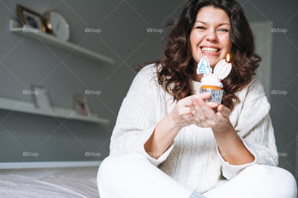Young adult happy beautiful woman forty years plus size body positive in white cozy knitted sweater with festive cupcake with candle in hands on bed at the home party, birthday girl