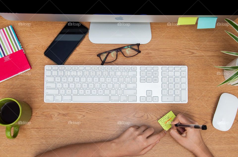 Desk seen from above. Woman’s hands writing on a post-it. 