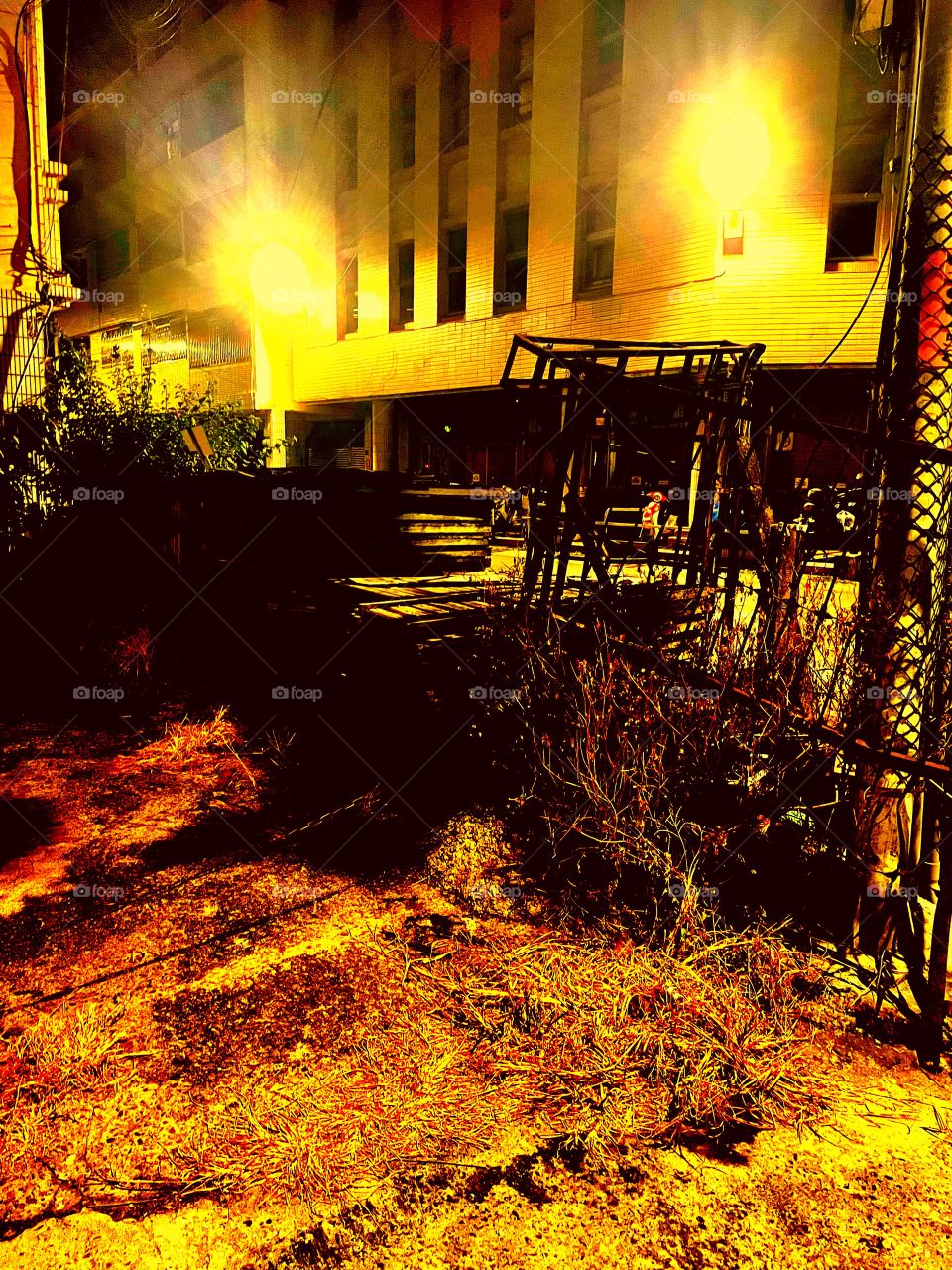 A junk site with several fall leaves on the ground. There are several crates in the background, and a tall rickety fence. In the background is an apartment building with two lights that look like angry eyes.