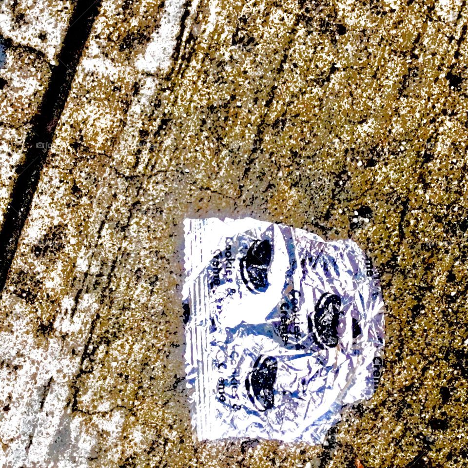 A little piece of silver aluminum foil in the shape of a mask or a human face on the asphalt of the sidewalk glowing golden in the evening sun gazes back at the observer. 2017, Bushwick, Bklyn. Hypnotic Productions