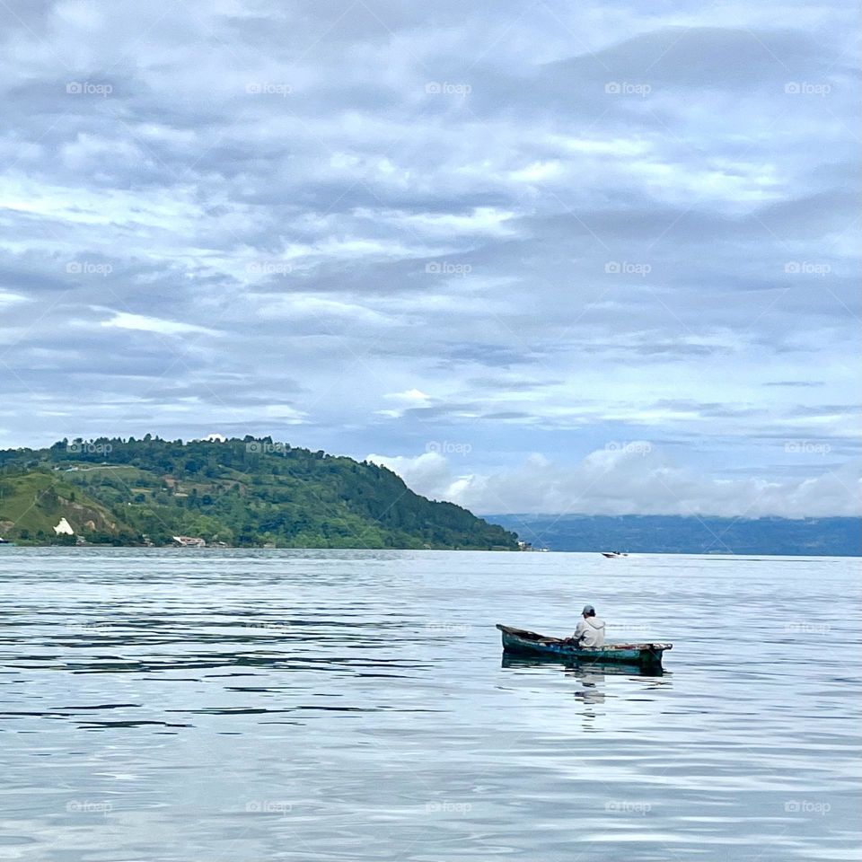 A fisherman is on his traditional boat while looking for some fish. The landscape is wide open, with sky touching the line of the lake.