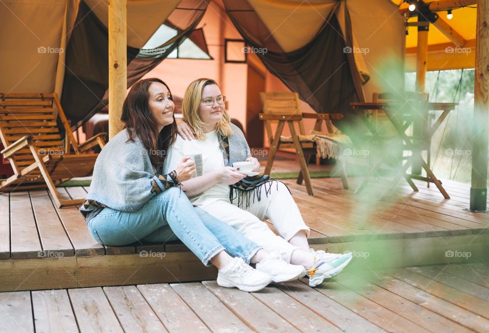 Two young women friends drinking tea and relaxing in glamping in the woods