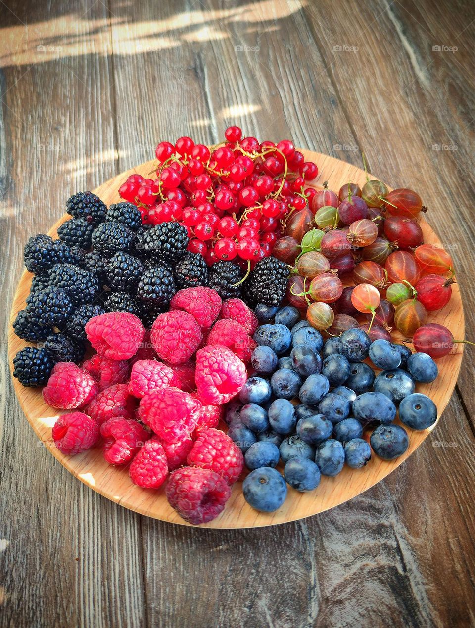 Summer treats. Five kinds of berries: red raspberry; black blackberry; red currants; green gooseberries and black blueberries on a brown plate, which stands on wooden boards. The sunlight of the summer sun falls on the composition and on the boards.