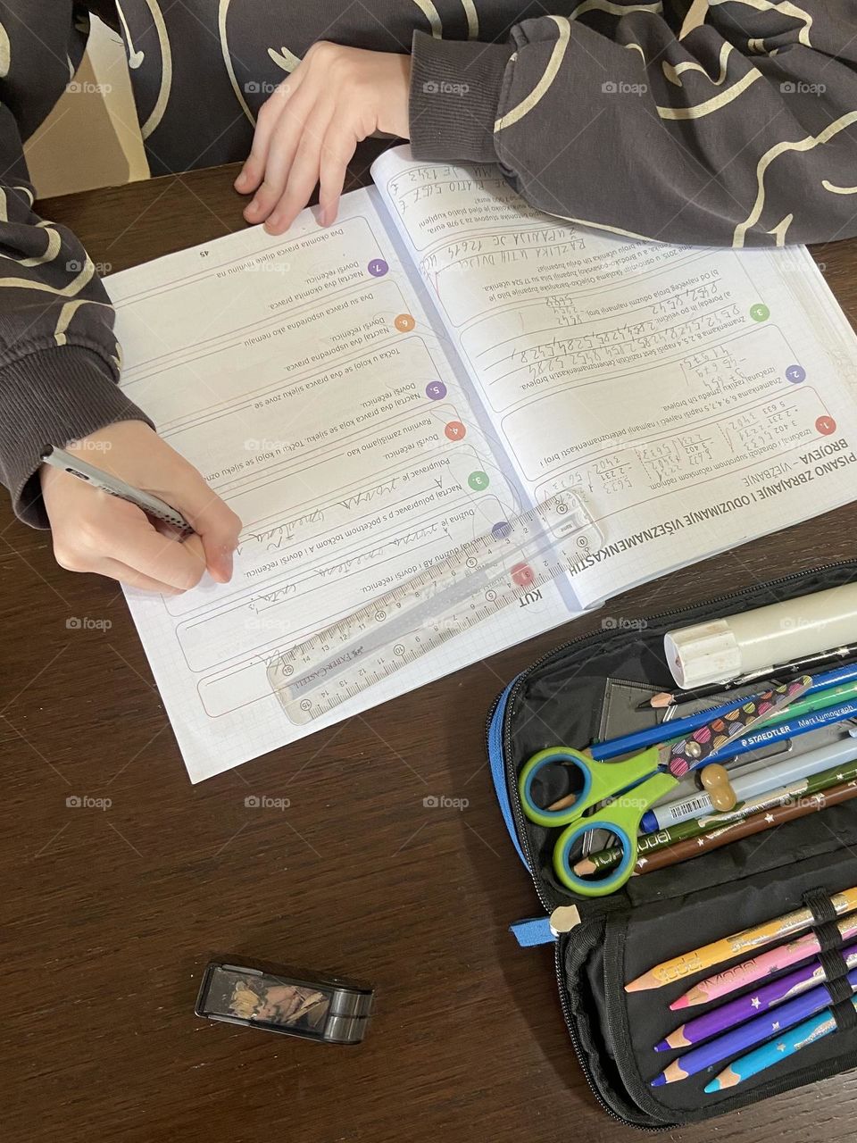 A young girl doing her homework work surrounded by her pencil case, a pencil sharpener and her ruler.